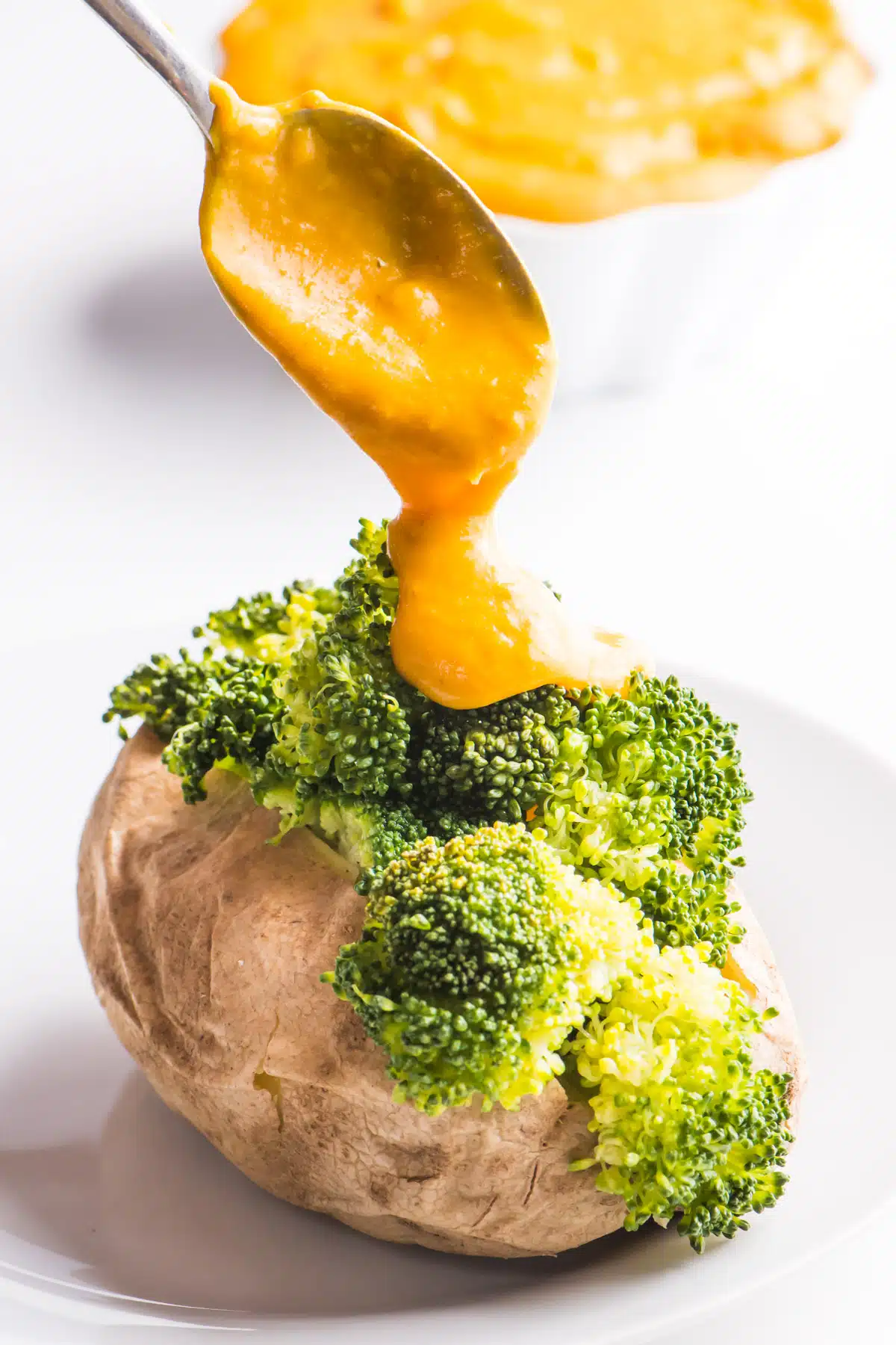 A spoon is drizzling vegan cheese sauce over a baked potato with steamed broccoli. 