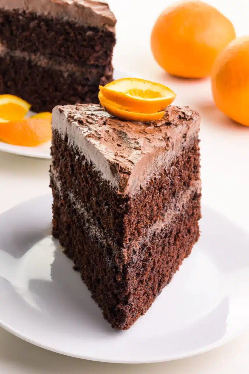 A slice of chocolate cake on a plate has an orange slice on top. There is another cake slice in the background and two oranges.