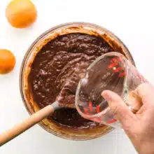 A hand holds a pyrex measuring cup, pouring hot water into chocolate batter in a mixing bowl. There is a spoon in the mixing bowl and two oranges sit next to the bowl.