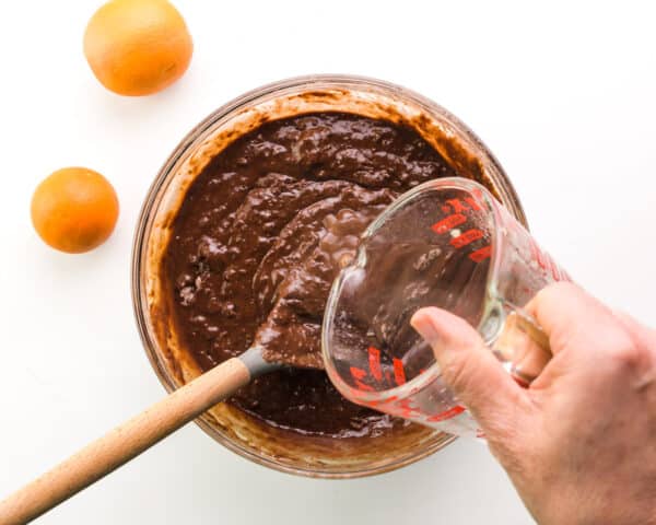 A hand holds a pyrex measuring cup, pouring hot water into chocolate batter in a mixing bowl. There is a spoon in the mixing bowl and two oranges sit next to the bowl.