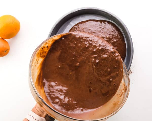 Chocolate cake batter is being poured into a round cake pan. There are oranges barely visible by the pan.