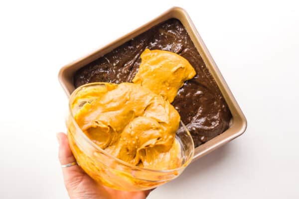 A hand holds a bowl, pouring pumpkin batter over brownie batter in a pan.