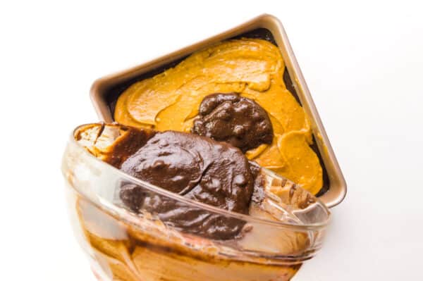 Brownie batter is being poured over a pumpkin batter layer in a baking pan.