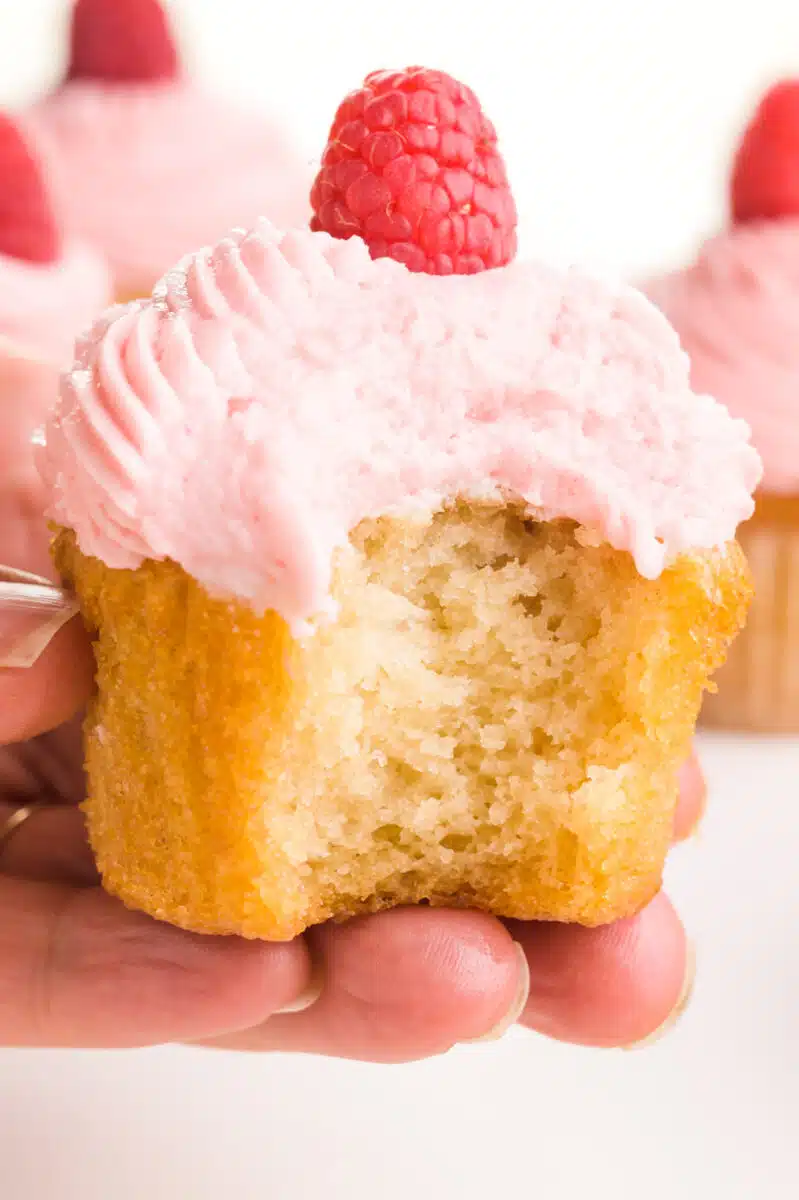 A hand holds a vanilla cupcake with a bite taken out. There is pink frosting on top and a fresh raspberry. There are other cupcakes visible in the background.