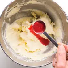 A hand holds a spoonful of mashed raspberries, pouring it over whipped butter in a mixing bowl.