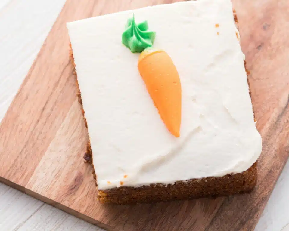 looking down on a slice of carrot cake on a cutting board. There is a frosting carrot on top of the cake slice.