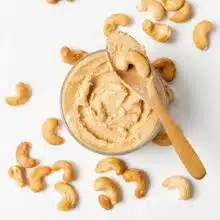 Looking down on a jar of cashew butter. There is a wooden spoon with cashew butter and a cashew. There are several cashews around the jar.