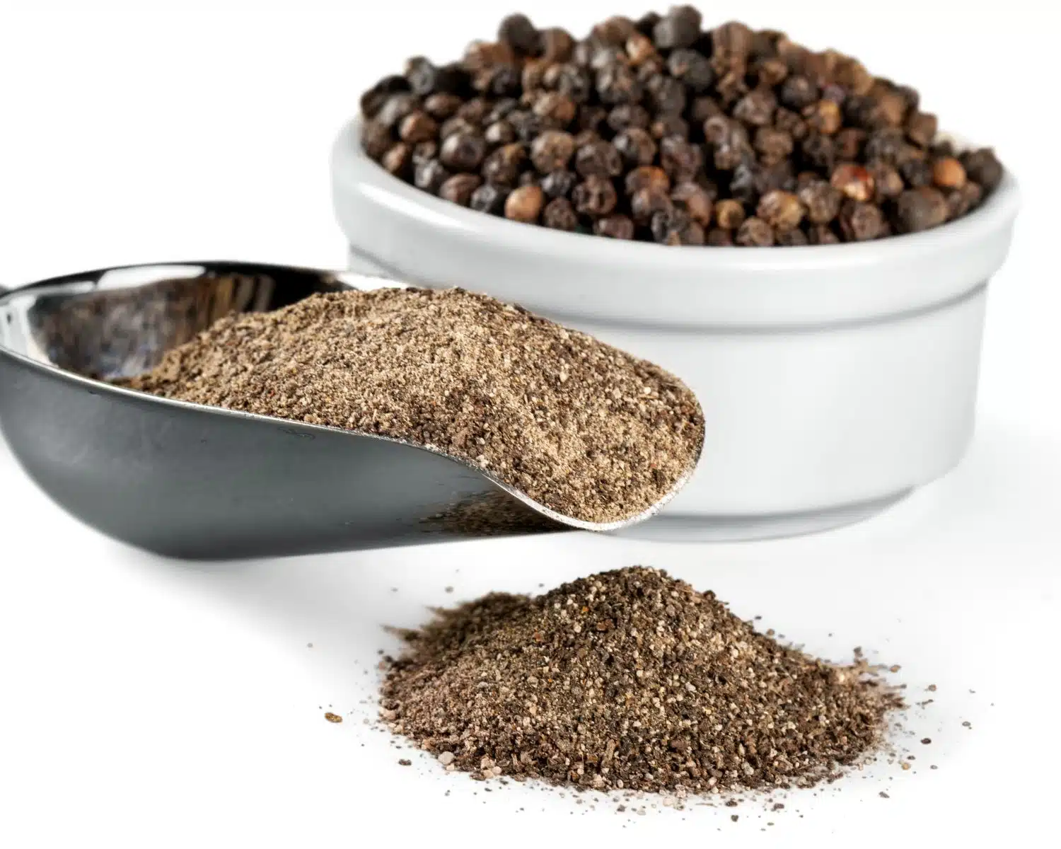 Peppercorns are in a bowl and a large scoop has freshly ground black pepper and some of it is scattered in front of the scoop.