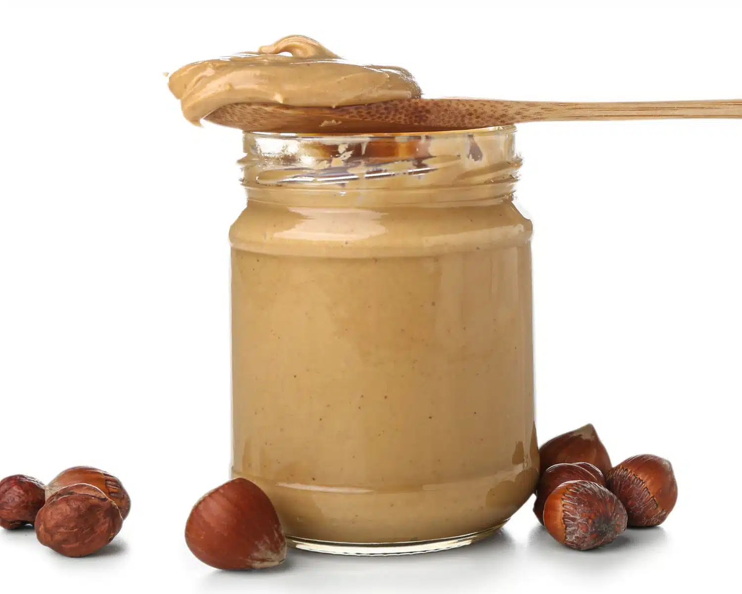 A spoon with nut butter sits on top of a jar of the same nut butter. There are hazelnuts around the jar.