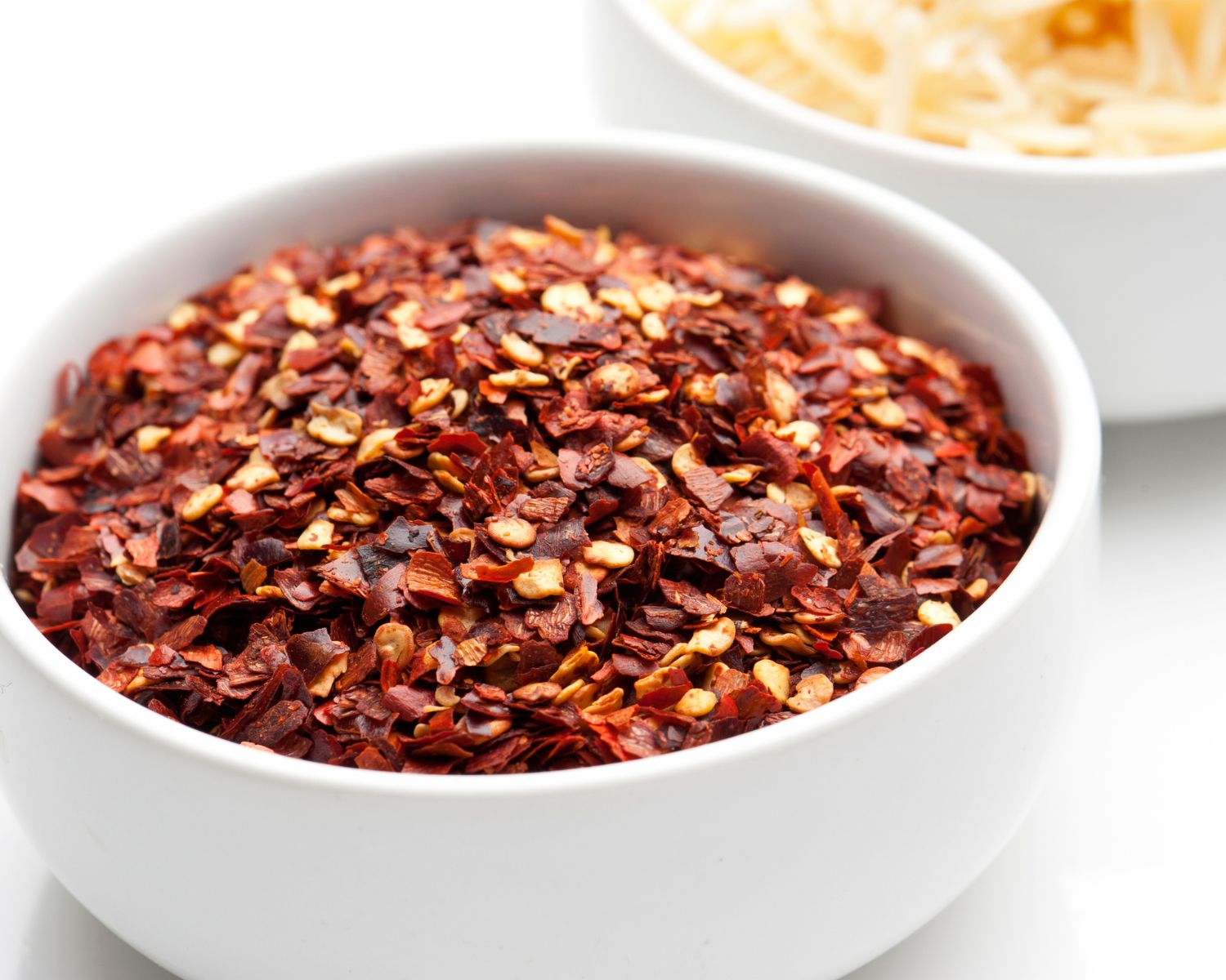 Red pepper flakes are in a bowl in front of a bowl of cheese shreds.