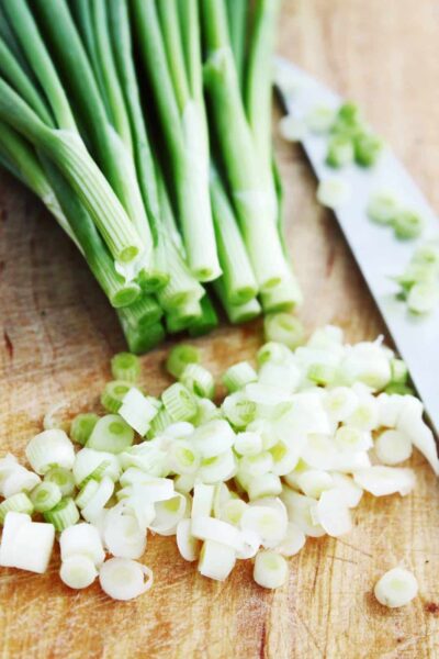 A bunch of spring onions are on a cutting board with some of the white parts of the onion finely chopped. There is a knife beside the onions.
