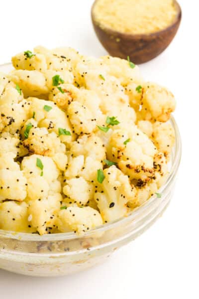 A bowl of air fryer cauliflower has fresh herbs on top. There is a bowl of nutritional yeast flakes in the background.