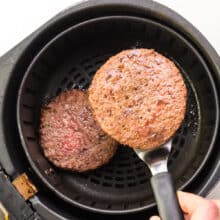 A hand holds a spatula with a veggie burger on it, preparing to flip it. It hovers over an air fryer basket.
