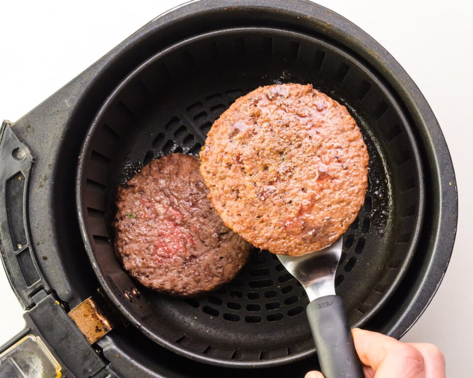 A hand holds a spatula with a veggie burger on it, preparing to flip it. It hovers over an air fryer basket.