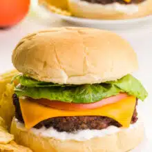 A cheeseburger is on a bun with other toppings, such as mayo, tomato, and greens. It sits on a plate with potato chips. There is another burger in the background along with a sliced tomato.