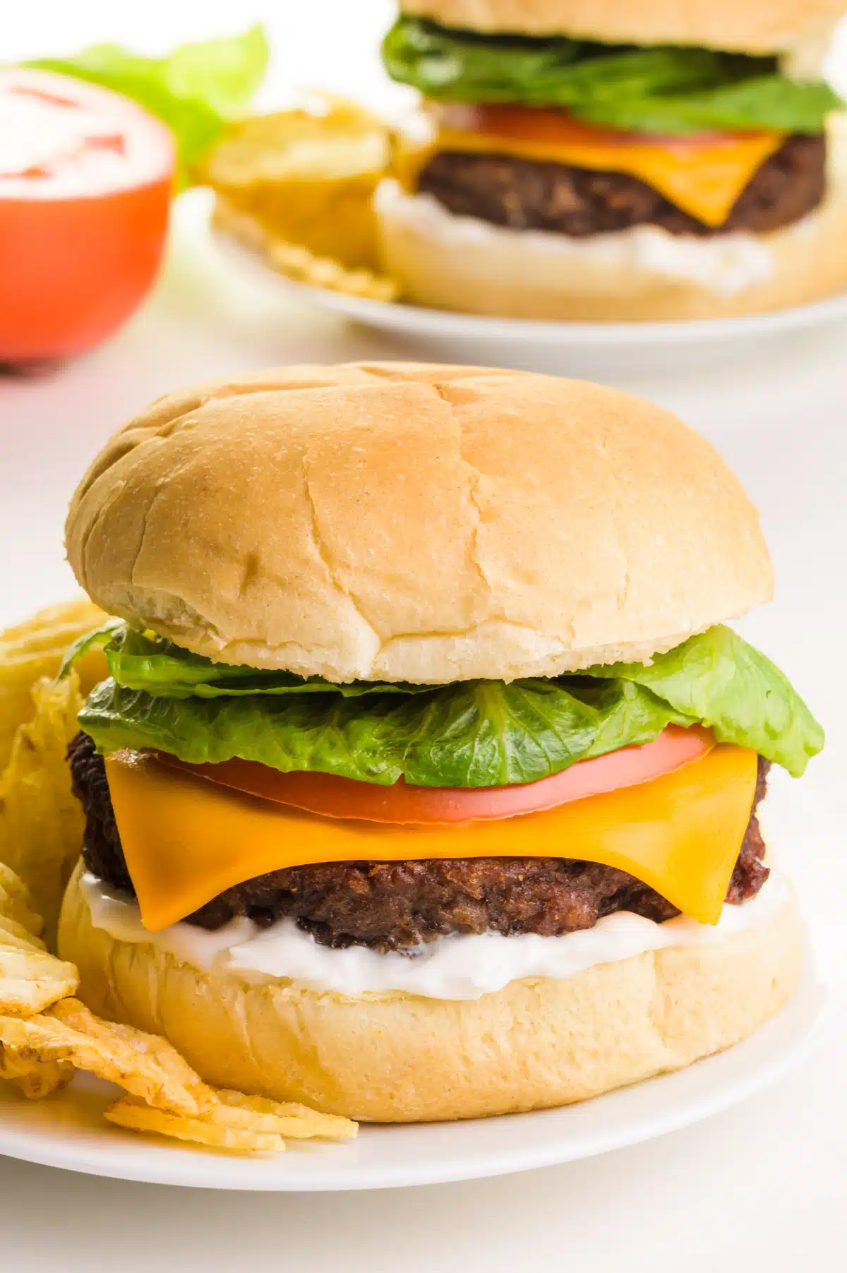 A cheeseburger is on a bun with other toppings such as mayo, tomato and greens.  It sits on a plate with potato chips.  In the background is another burger with a slice of tomato.