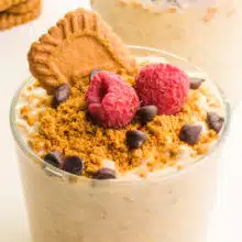 A jar of biscoff overnight oats has crumbled cookies on top with raspberries, chocolate chips, and a Biscoff cookie.