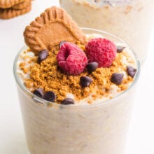 A jar of biscoff overnight oats has crumbled cookies on top with raspberries, chocolate chips, and a Biscoff cookie.