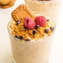 A glass jar of overnight oats has a Biscoff cookie on top with raspberries and chocolate chips. There is another jar of the oats in the background sitting beside more cookies.