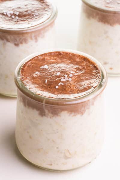 A bowl of bounty overnight oats has a chocolate topping and flaky sea salt on top. There are two jars of the same oats in the background.