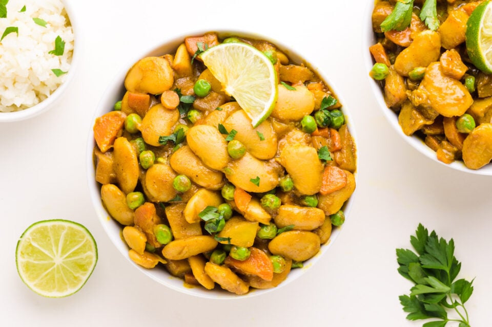 Looking down on a bowl of butter bean curry with a lime wedge on top. There is another bowl next to it, along with fresh limes, rice, and fresh herbs.