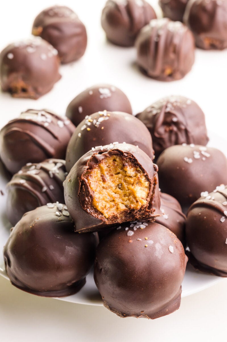Cashew butter truffles are on a plate, one with a bite taken out. There are more of the treats in the background.