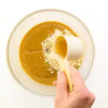 Cashew butter is in a bowl with other ingredients. A hand pours syrup sweetener from a measuring cup into the bowl.