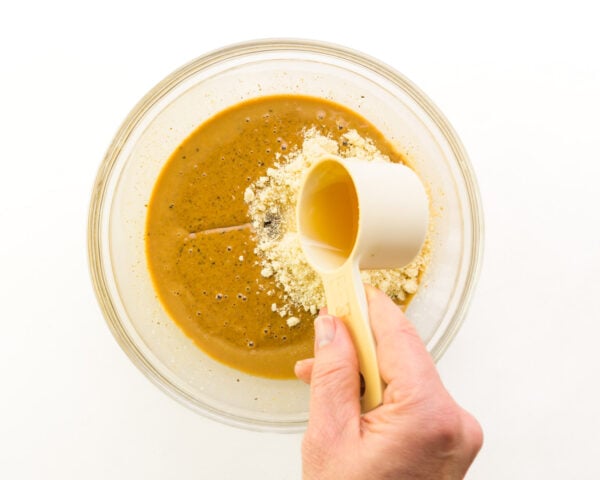 Cashew butter is in a bowl with other ingredients. A hand pours syrup sweetener from a measuring cup into the bowl.