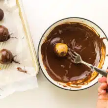A cashew butter ball is being dipped in a bowl with melted chocolate. There are freshly dipped balls on a tray next to the bowl.
