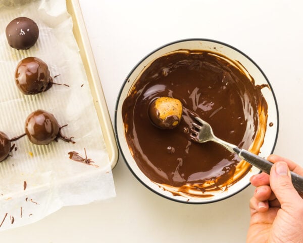 A cashew butter ball is being dipped in a bowl with melted chocolate. There are freshly dipped balls on a tray next to the bowl.