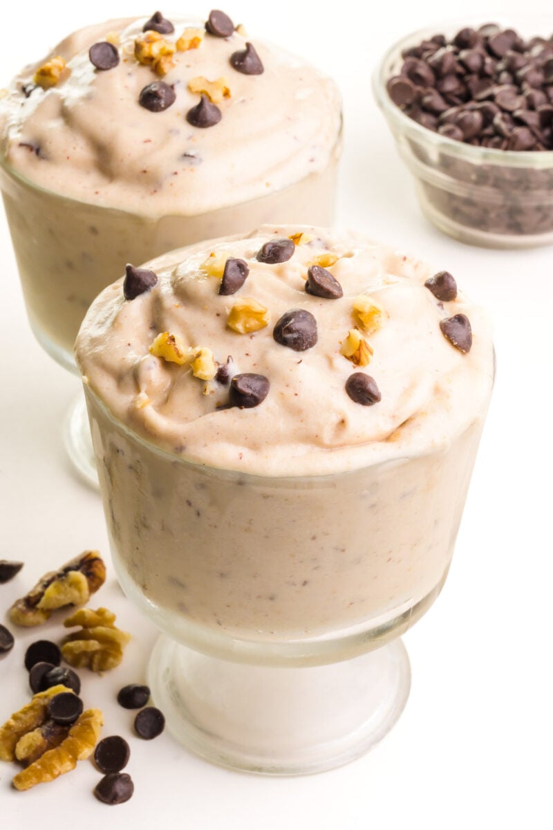 Two bowls contain vegan chunky monkey ice cream with chocolate chips and walnuts on top. There are chocolate chips and walnuts next to the serving dishes and a bowl in the background as well.