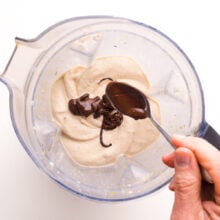 A hand holds a spoon, drizzling melted chocolate into a blender with whipped bananas.