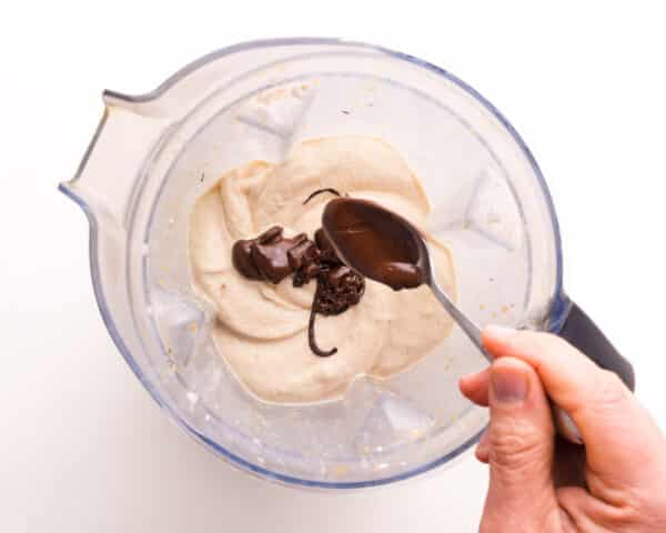 A hand holds a spoon, drizzling melted chocolate into a blender with whipped bananas.