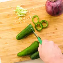 Looking down on a hand holding a paring knife, cutting a jalapeño. There are more jalapeños, the pith and seeds, and a red onion on the cutting board.