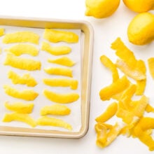 Several strips of lemon zest on a baking sheet. There are pieces of lemon zest around the pan along with fresh lemons.