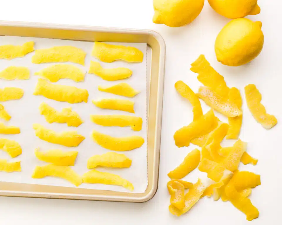 Several strips of lemon zest on a baking sheet. There are pieces of lemon zest around the pan along with fresh lemons.