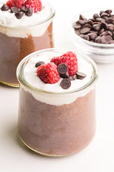 A small glass jar holds chocolate flax pudding topped with creamy white yogurt, chocolate chips an raspberries. There is another jar and a bowl of chocolate chips in the background.