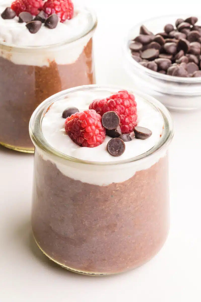 A small glass jar contains chocolate flax pudding topped with creamy white yogurt, chocolate chips and raspberries.  In the background is another jar and a bowl of chocolate chips.