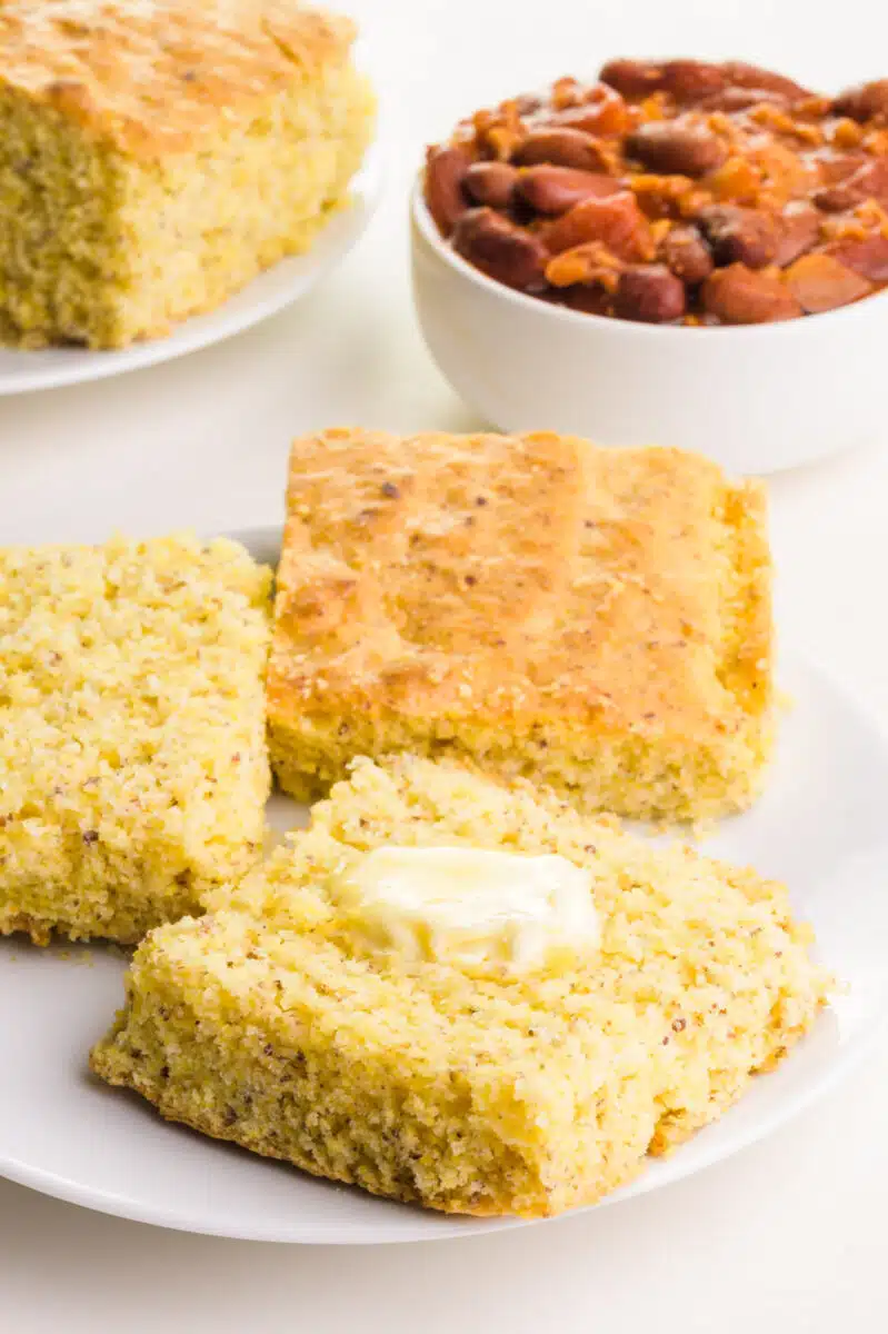 Cornbread slices are sitting on a plate, one with a pat of butter on top. There is a bowl of chili and more cornbread in the background.