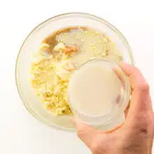 A hand holds a bowl of soy milk, pouring it into a bowl with cornbread ingredients.