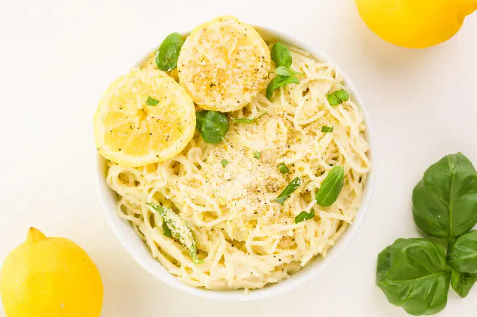 Looking down on vegan lemon pasta in a bowl with fresh lemon slices and basil. There are basil and lemons near the bowl.