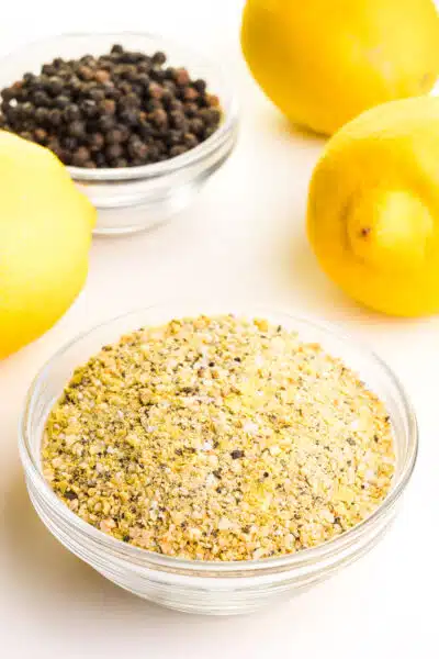 A bowl of lemon seasoning has fresh lemon and a bowl of black peppercorns behind it.