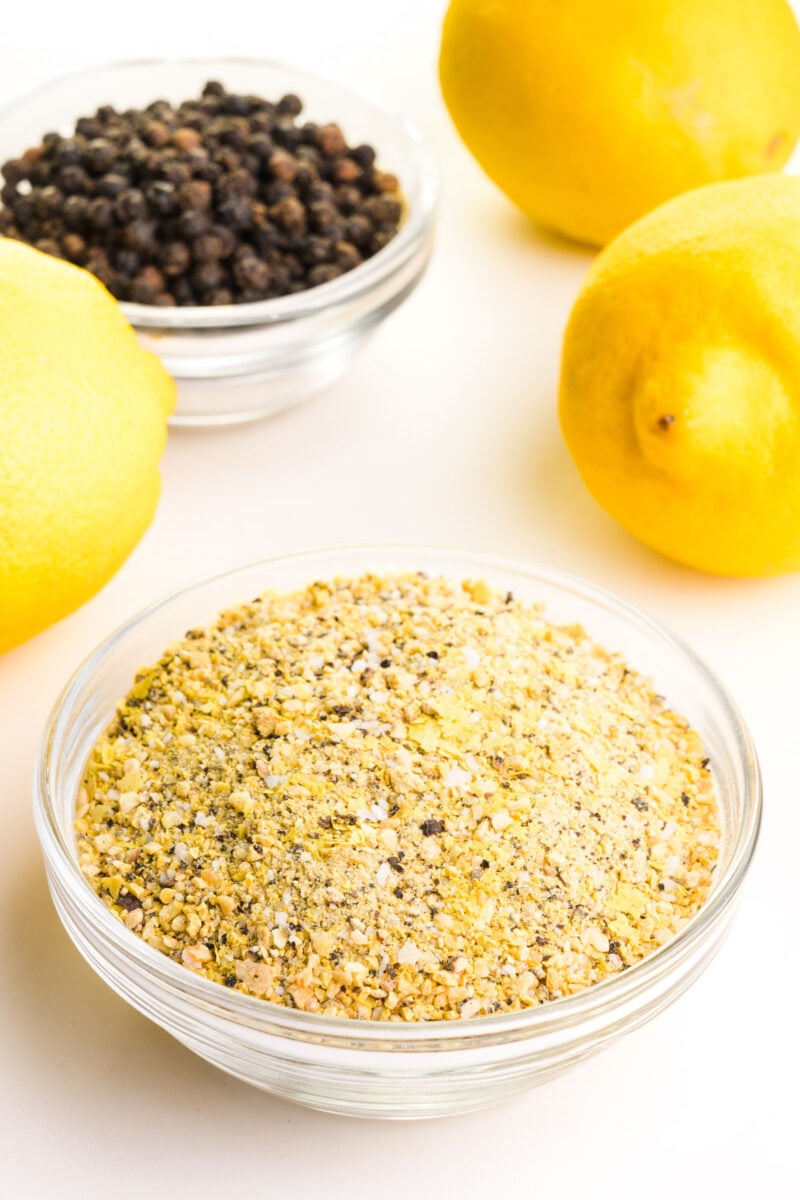 A bowl of lemon seasoning has fresh lemon and a bowl of black peppercorns behind it.