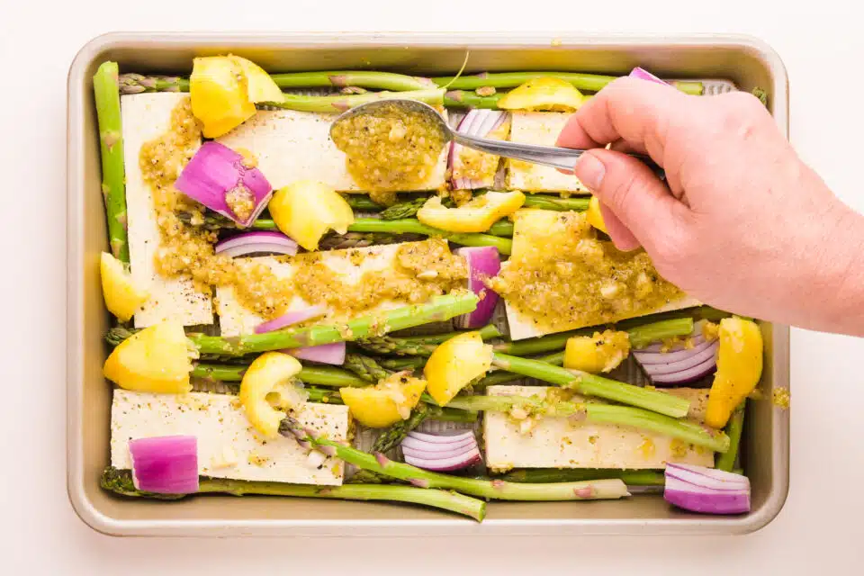 A hand spoons extra marinade on tofu in a baking pan with red onions, lemons, and asparagus.