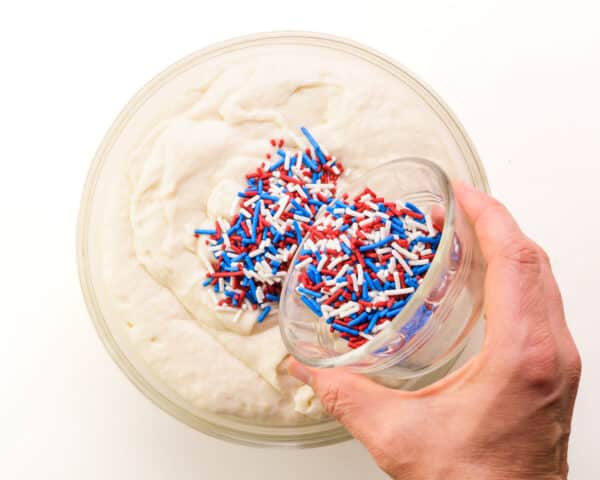 A hand holds red, white, and blue jimmies sprinkles, pouring them into a bowl with vanilla cake mix batter.