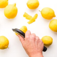 A hand holds a zester, removing the skin from a fresh lemon. There are several fresh lemons around it.