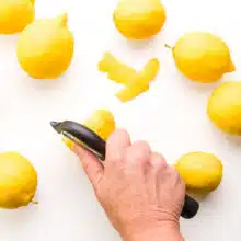 A hand holds a zester, removing the skin from a fresh lemon. There are several fresh lemons around it.