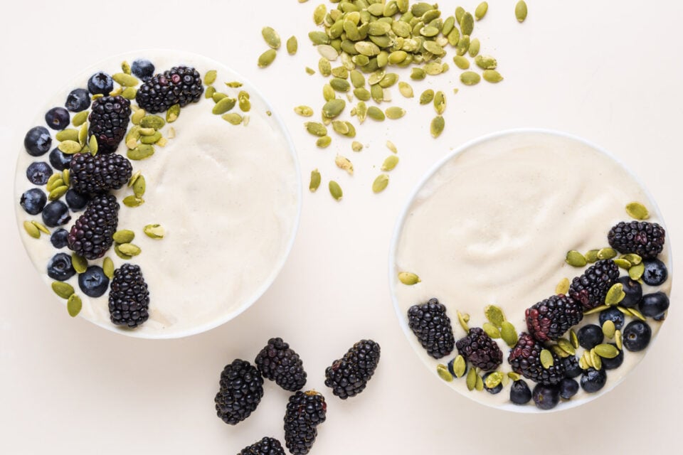 Looking down on two vanilla smoothie bowls with fresh berries and pumpkin seeds on top. There are more toppings between the bowls.