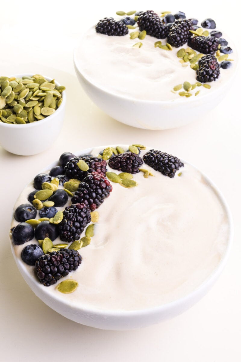 Two bowls of vanilla smoothie has berries and seeds on top. There's a bowl of pumpkin seeds in the background.