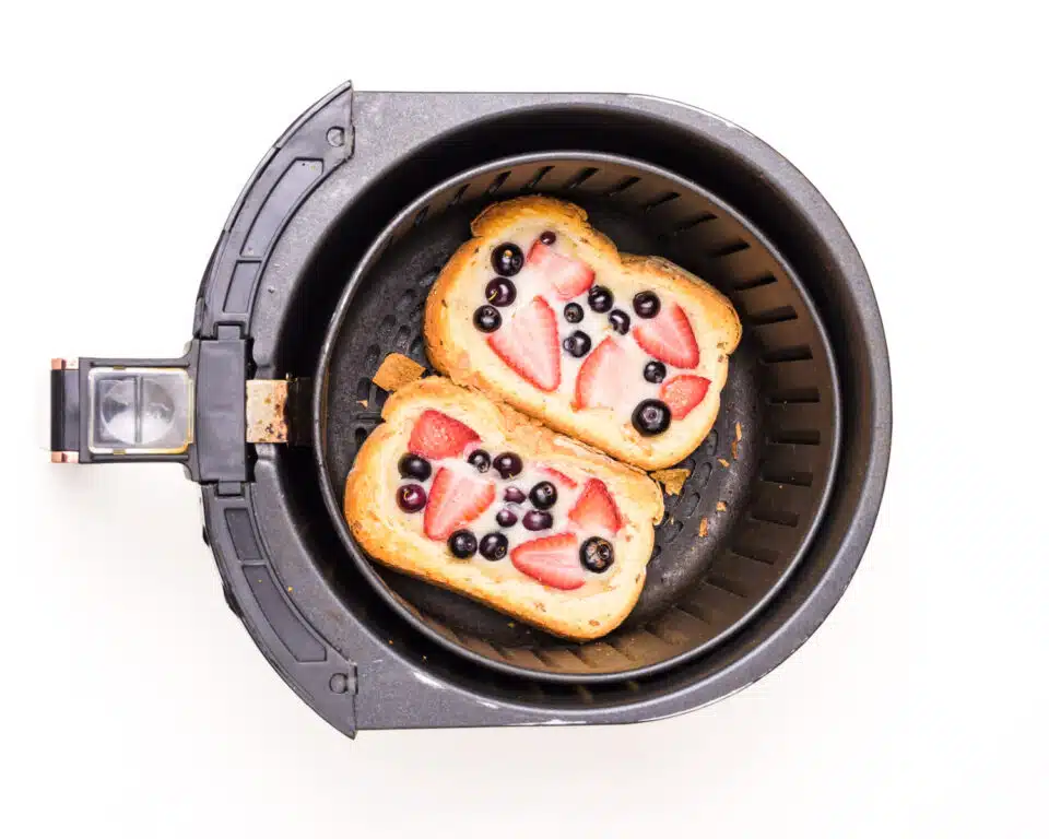 An air fryer basket holds two freshly cooked slices of eggless yogurt toast with berries on top.